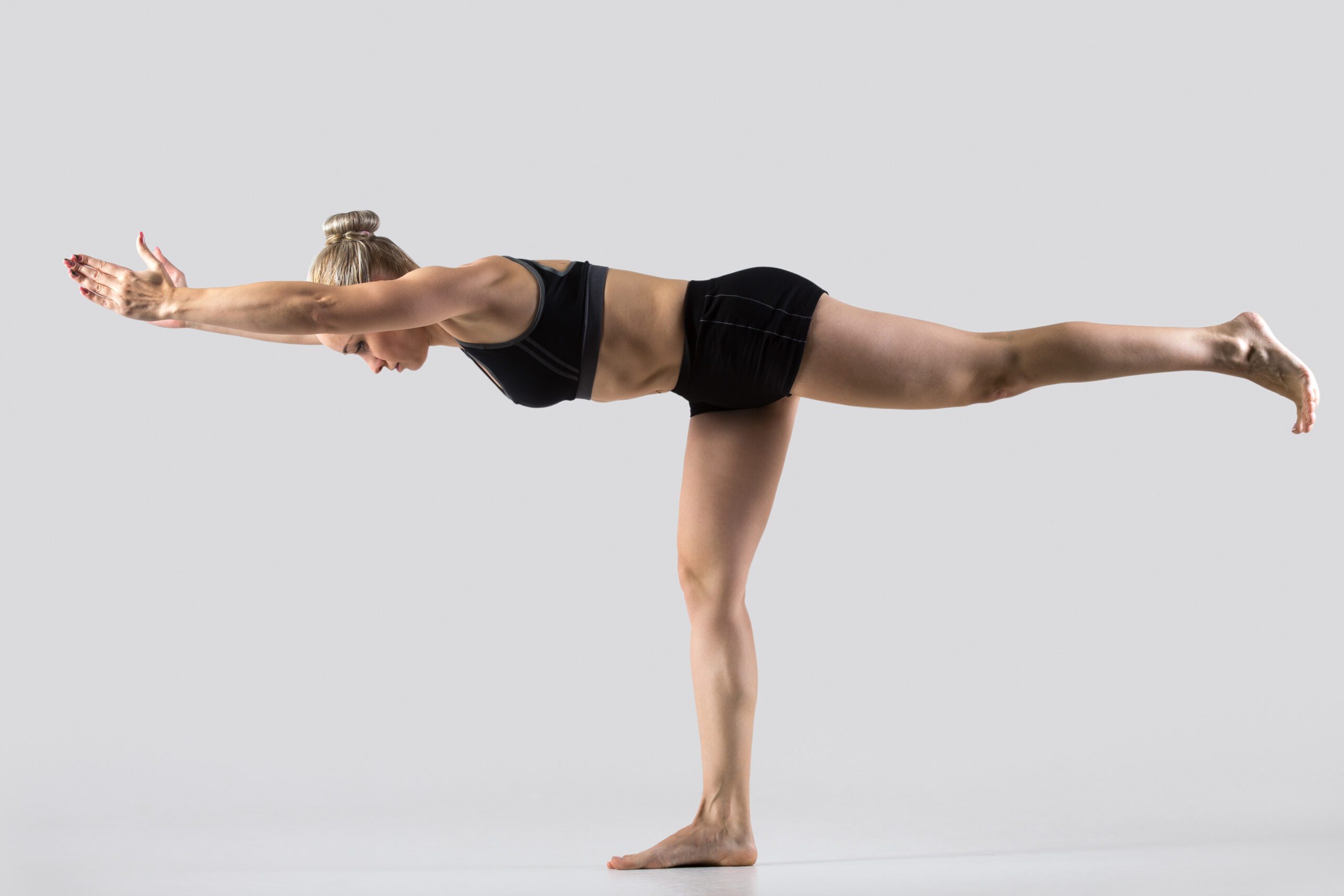 Sporty beautiful young woman practicing yoga, standing in Warrior III posture (Virabhadrasana 3), working out wearing black sportswear, studio full length, grey background